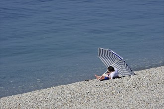 Tourist reading book under sunshade on pebble beach at Etretat, Côte d'Albâtre, Upper Normandy,