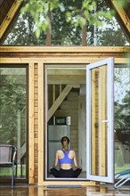 Back view of young woman sitting in lotus yoga pose inside wooden cabin in front of open door