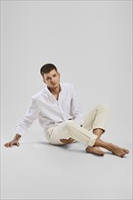 Full length studio portrait of young barefoot man in white shirt and ivory trousers sitting the