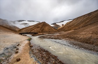 Bridge over steaming stream between colourful rhyolite mountains with snowfields, Hveradalir