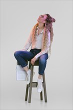 Woman with dreadlocks sits on tall chair, holds magazine in hand and looks up