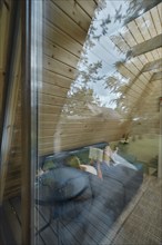 View through the window at young woman on sofa inside summer house reading a book