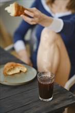 Unrecognizable woman sitting in the kitchen with croissant in hand, focus on glass with coffee