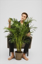 Barefoot young man in black suit siting in an armchair and looking at home plant in the studio