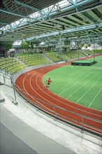 Person jogging in a stadium on a red running track, green pitch and yellow stands, covered area,