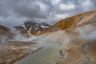 Steaming stream between colourful rhyolite mountains with snowfields, Hveradalir geothermal area,