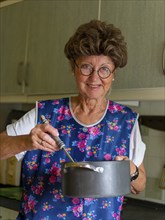 Granny with old smock apron, glasses and wig in the kitchen, holding cooking pot and ladle,