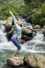 Young sporty fit woman doing yoga asana Utkatasana (chair pose) outdoors at tropical waterfall