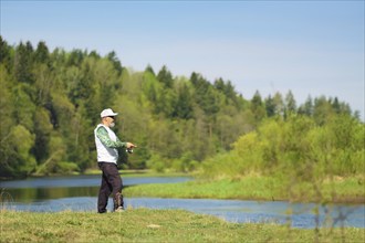 Fisherman with a spinning rod catching fish on a river at sunny summer day with green trees at