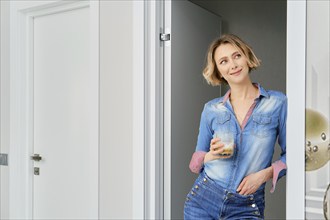 Young woman stands in the doorway of the bedroom with a glass of cappuccino in her hand