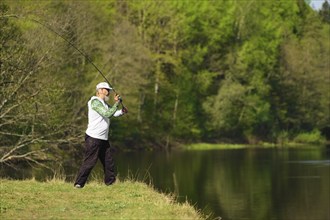 Fisherman with a spinning rod catching fish on a river at sunny summer day with green trees at