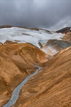 Steaming stream between colourful rhyolite mountains with snowfields, Hveradalir geothermal area,