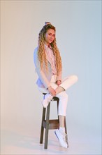 Young woman with dreadlocks in jeans and jacket sits on chair under colorful lights