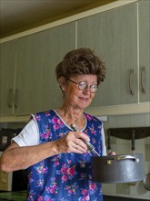 Granny with old smock apron, glasses and wig in the kitchen, holding cooking pot and ladle,