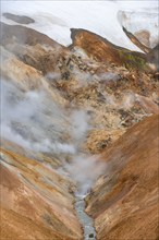 Steaming stream between colourful rhyolite mountains and snowfields, Hveradalir geothermal area,