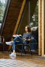 Cute woman sits in wicker chair at wooden terrace and reading the book after the rain