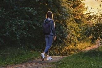 Back view of an active woman goes along path from forest to clearing in rays of sunset