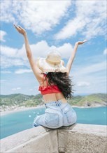 Lifestyle of woman on vacation sitting spreading hands to the beach. Tourist girl in hat spreading