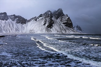 White foamy waves on the beach of Stokkness, behind snowy rocky slopes of Vestrahorn, near Höfn,