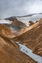 Steaming stream between colourful rhyolite mountains and snowfields, Hveradalir geothermal area,