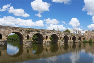 Ancient stone bridge with reflections in the river water, under blue sky with some clouds, Roman