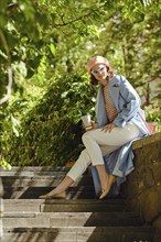 Hipster woman sitting at an outdoor stone bench, enjoying tea and sunshine