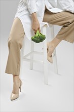 Daily healthy eating concept. Cropped image of woman sitting on chair with broccoli in hand