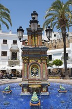 Decorative fountain with ceramic details and four lanterns, surrounded by palm trees, in warm