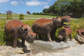 Family of metal sculptures depicting hippos on a stone foundation in a green park under a blue sky,