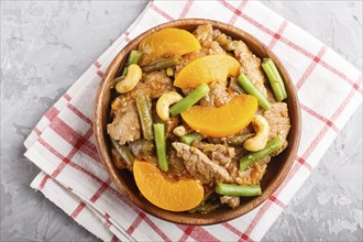 Fried pork with peaches, cashew and green beans in a wooden bowl on a gray concrete background. Top