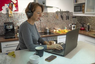 Woman, mid-50s, works from home, with laptop and communicates with colleagues via headset, home