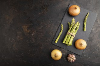 Bunch of fresh green asparagus, garlic, onion on slate board on black concrete background. Top