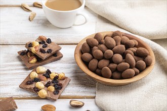 Almond in chocolate dragees in wooden plate and a cup of coffee on white wooden background and