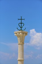 Stone column with the decorative cross of the Camargue, in front of a blue sky with clouds, summer,