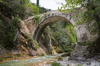 Old stone bridge spans a calm river surrounded by lush greenery, bridge of Platanias over the river