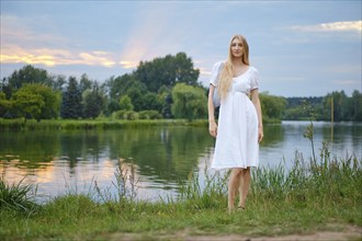 Young woman with long blonde hair standing on the river bank during sunset