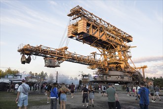 Festival visitors in front of an open-cast mining excavator illuminated by the evening sun at the