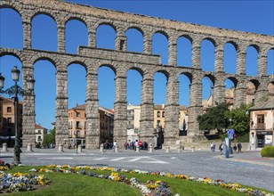 A historic aqueduct with stone arches spans a town with a flowerbed in the foreground under a blue