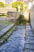 Picture of a small fountain with flowing water. Paving stones and greenery in the background,