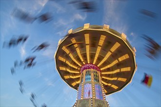 An illuminated carousel at dusk, spinning fast, in front of a blue sky with clouds and a Germany