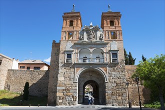 Historic gate with towers and city wall, people walking underneath, blue sky in the background,