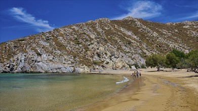 Golden sandy beach and crystal clear water with walkers, surrounded by hills and trees under a blue