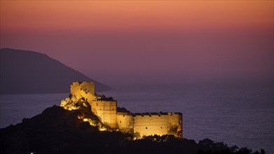 View of an illuminated castle ruin at dusk, surrounded by sea and mountains under a colourful sky,