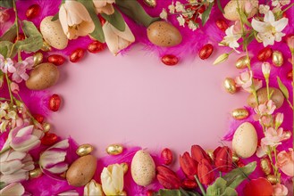 Frame of eggs and spring flowers on pink feathers with red and gold chocolate eggs on a pink