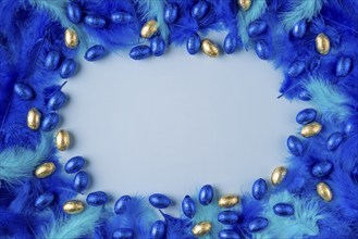 Frame made of blue and turquoise feathers with blue and gold chocolate eggs on a light blue