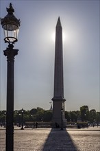 An obelisk rises behind a lantern, sunlight intensifies the silhouettes, Paris