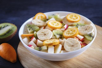 Vegetarian salad of bananas, apples, pears, kumquats and kiwi on black woodem background, close up,