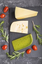 Green basil cheese and various types of cheese with rosemary and tomatoes on black slate board on a