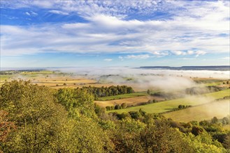 Beautiful view at a rural landscape plain with fields and morning mist from a hill with trees in