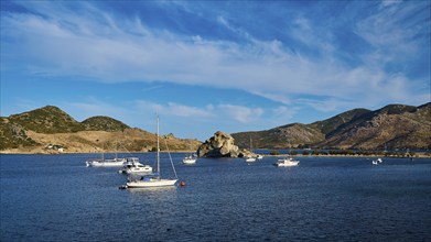 Boats on calm blue water, surrounded by mountains and rocks under a cloudy sky, Petras Beach,